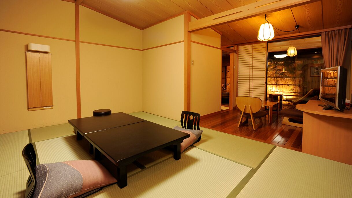 Una tradizionale stanza giapponese con pavimento in tatami, un tavolino, sedie, una televisione e una porta scorrevole che dà su un'altra stanza.