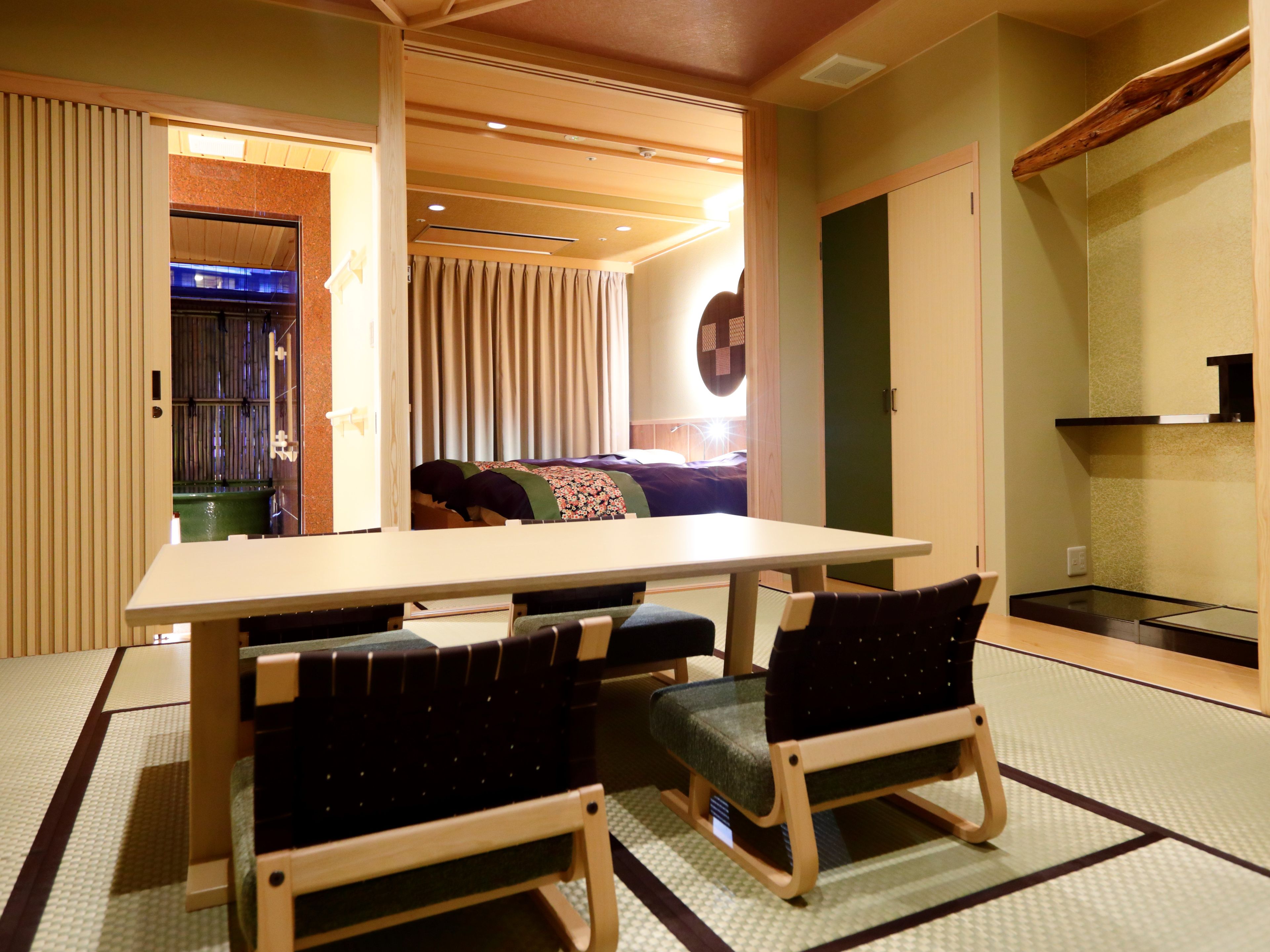 A room with tatami flooring, a low table, two chairs, a sofa, and a sliding door leading to a bathroom.