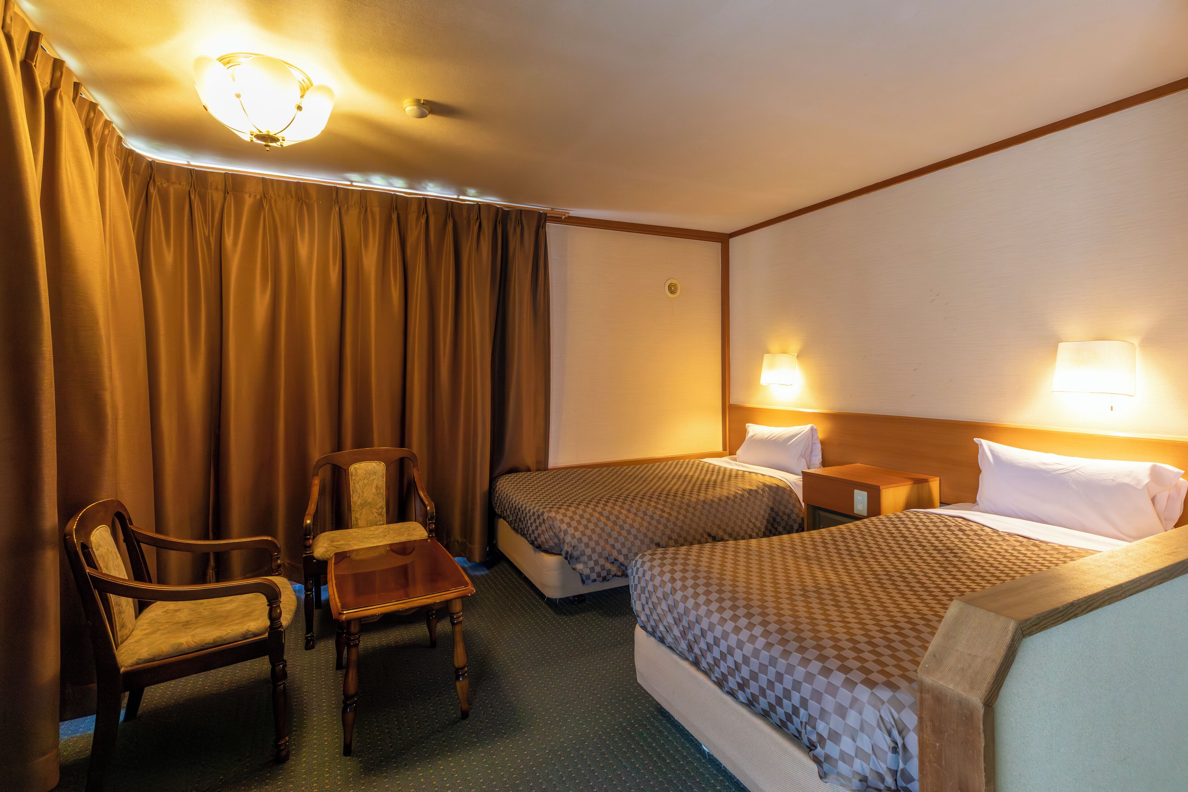 A hotel room with two beds, a desk, two chairs, and a nightstand with lamps.