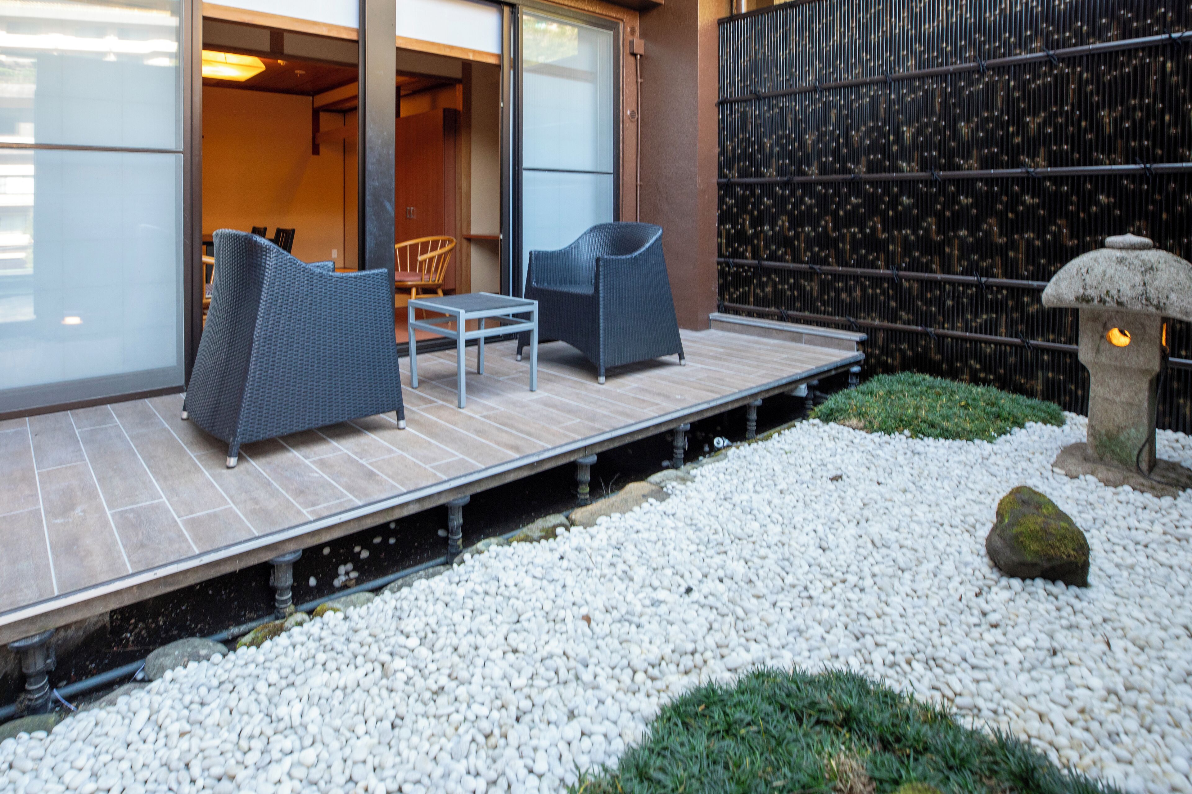 An outdoor seating area with two chairs and a table, a stone lantern, and a pebbled garden path.
