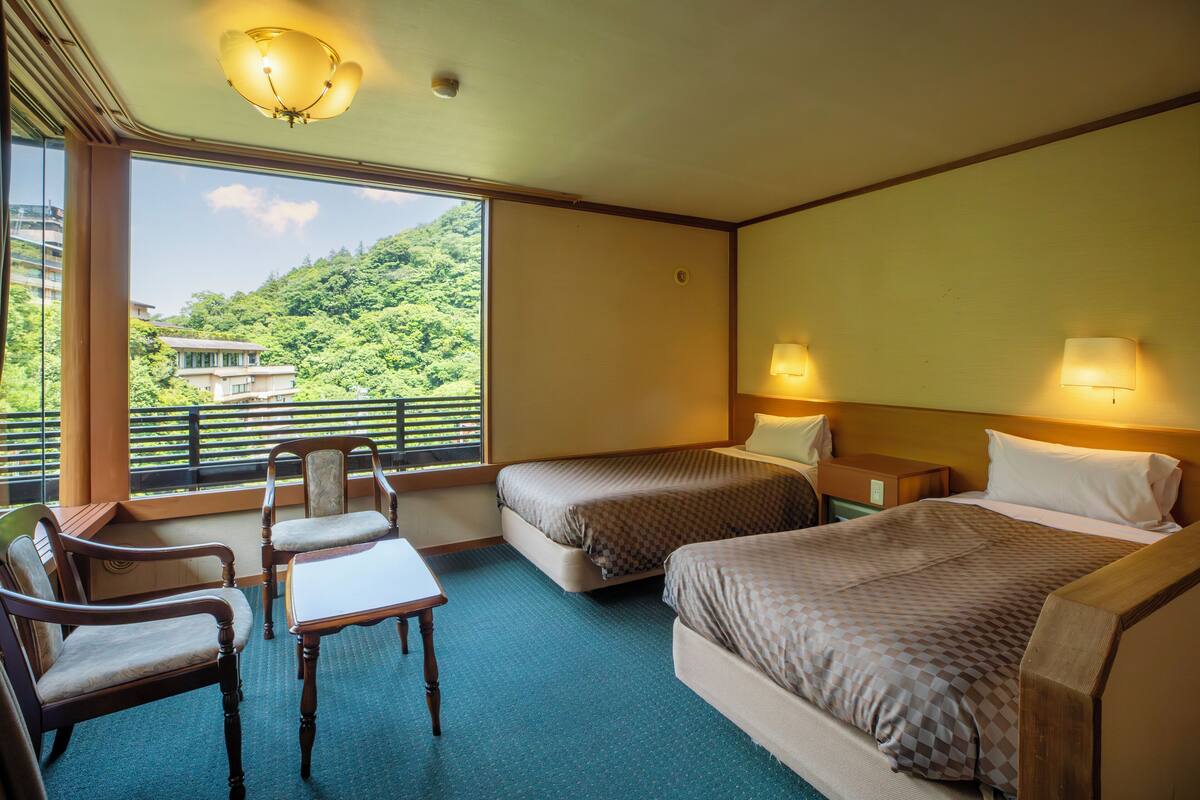 A hotel room with two beds, a desk, chairs, and a balcony with a view of a hillside.