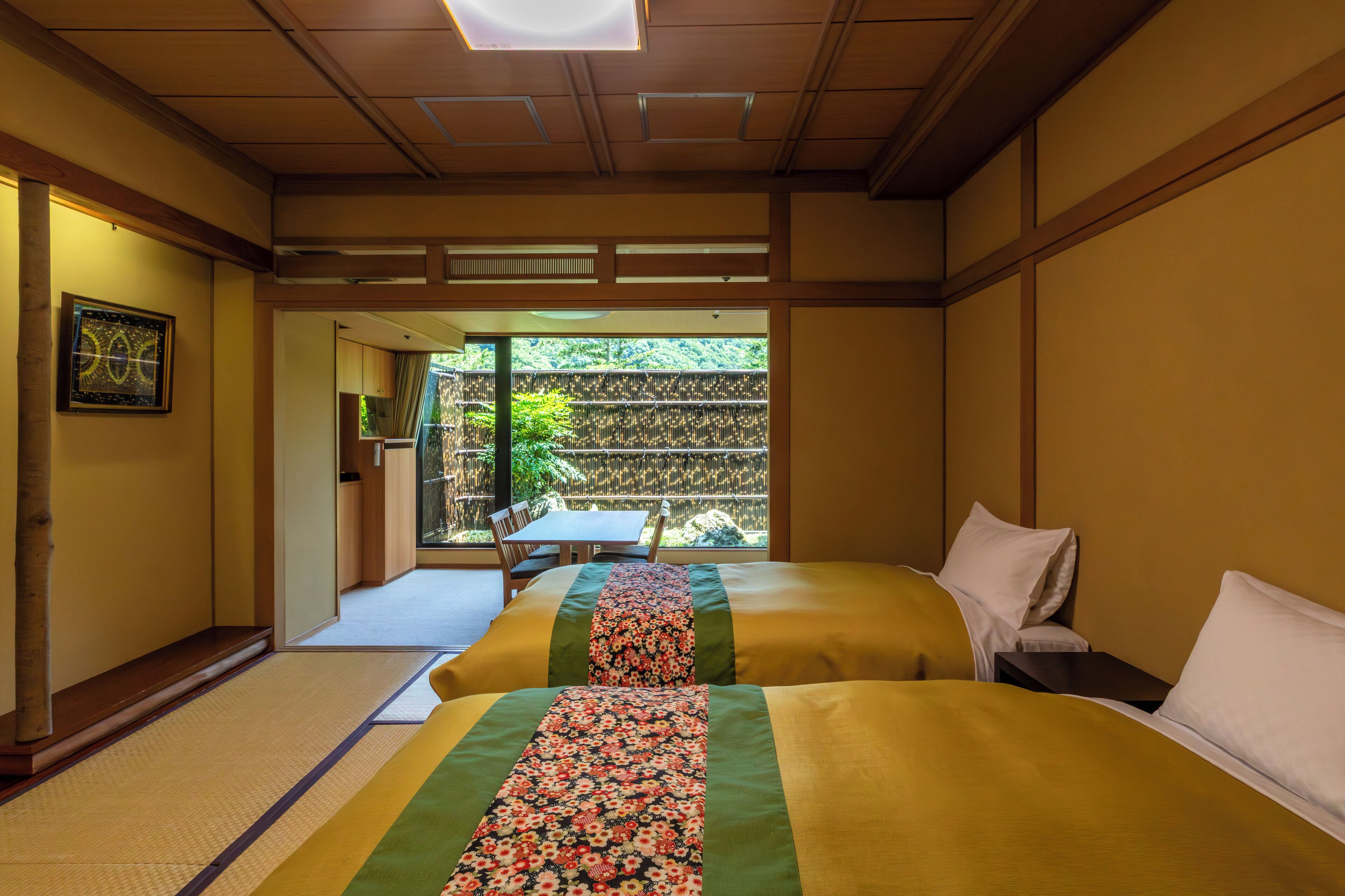 Ein traditioneller japanischer Raum mit Tatami-Böden, zwei Betten mit floralen Tagesdecken, einem Holztisch und Blick auf einen Bambuszaun und Grünflächen vor dem Fenster.