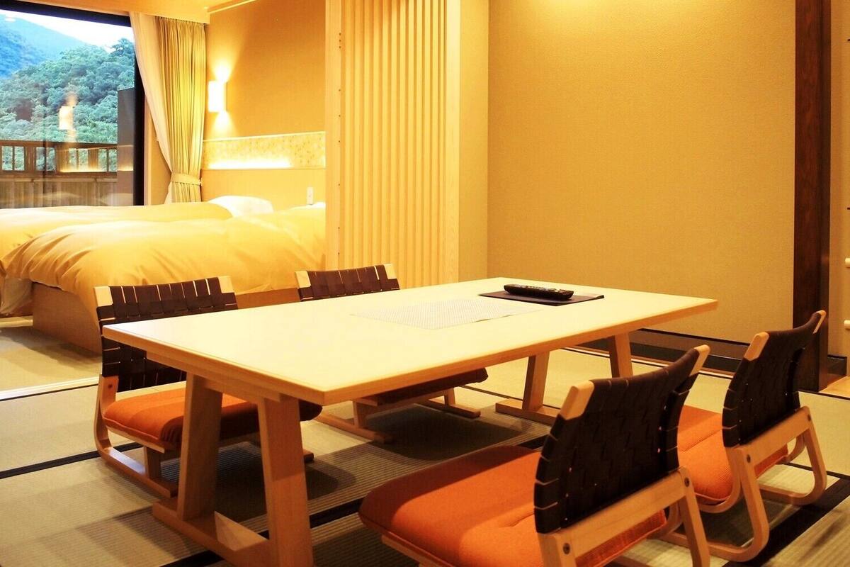 A hotel room with a bed, a dining table with chairs, and a view of a forest.