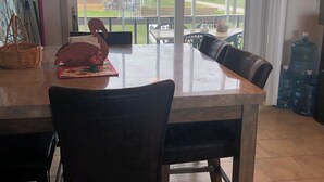 Un comedor con una mesa grande y sillas, vistas a una terraza exterior a través de un ventanal, y un dispensador de agua al fondo.