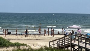 Una playa con un paseo de madera que conduce al agua, gente jugando al voleibol y una bandera a la derecha.