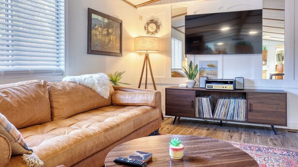 A living room with a leather sofa, a coffee table with a remote control and a plant, a floor lamp, a TV, and a bookshelf with CDs. - Rainier Bunkhouse & Bar-Cade- HotTub,MtnView,2 blks to Packwood Brewing! Pets ok (Packwood)