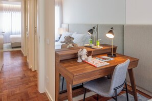 A hotel room with a wooden desk, a chair, a bed with white bedding, a teddy bear, a lamp, and a nightstand with a mirror.
