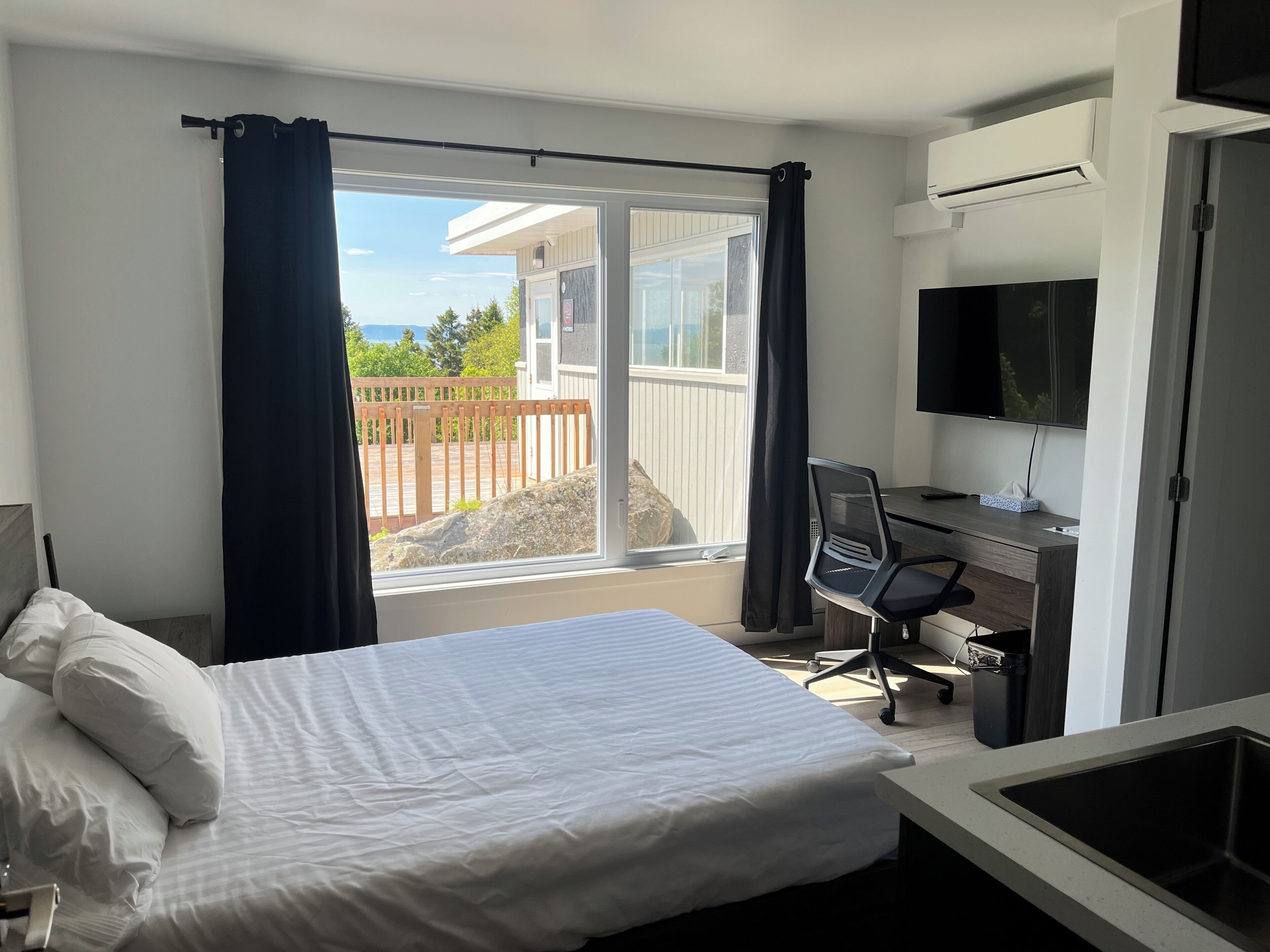 A hotel room with a bed, a desk with a chair, a television, and a view of a balcony with a wooden deck and a building in the background.