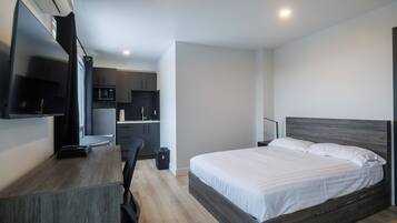 A modern hotel room with a wooden bed, a desk with a flat-screen TV, a kitchenette, and a large window.