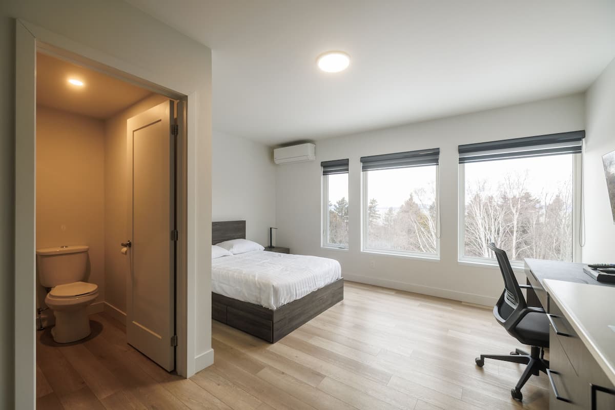 A bedroom with a bed, a desk, and a chair. There are three windows and an air conditioning unit on the wall.