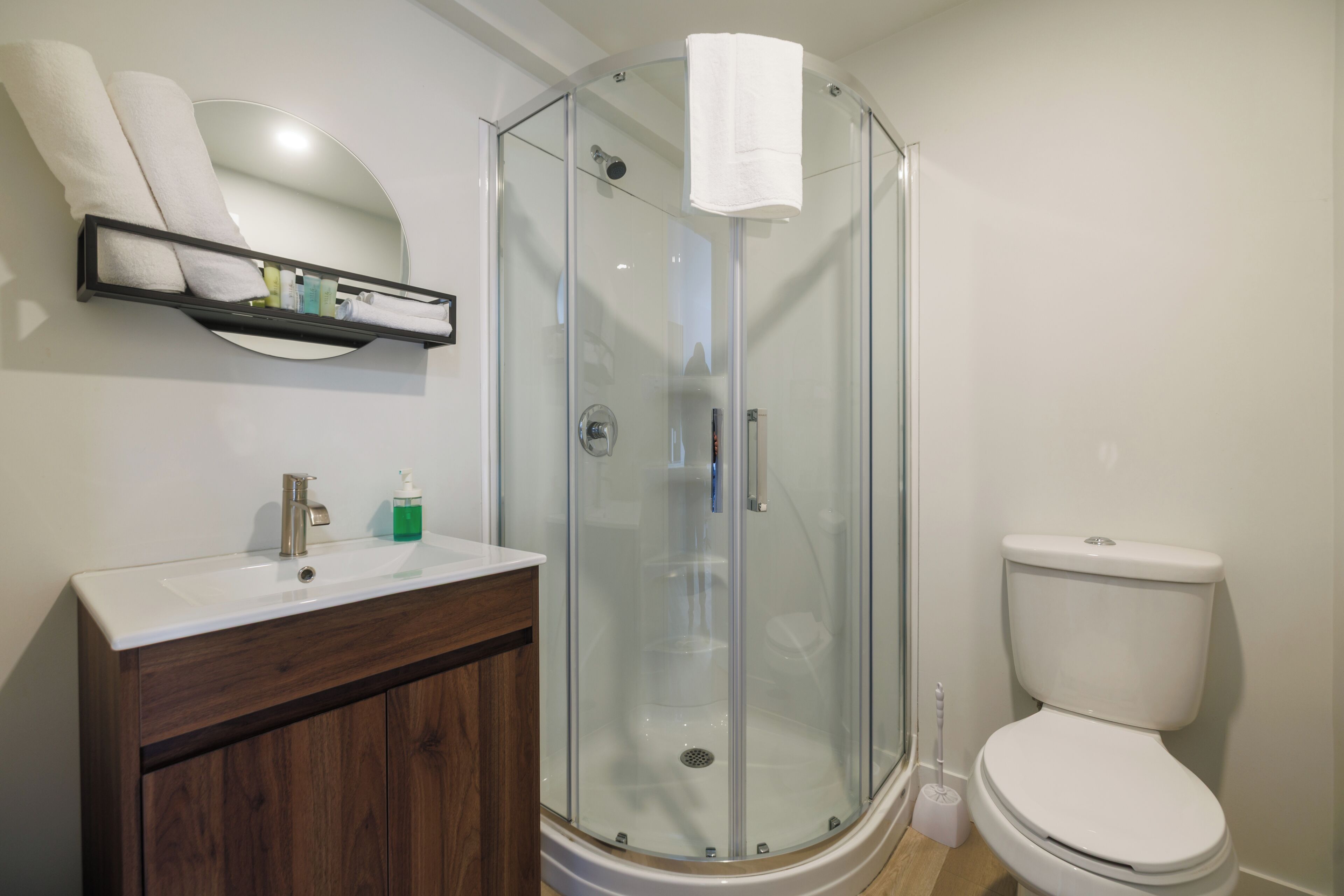 Un baño con cabina de ducha de vidrio, un inodoro blanco, un tocador de madera con un espejo y un estante con artículos de aseo.