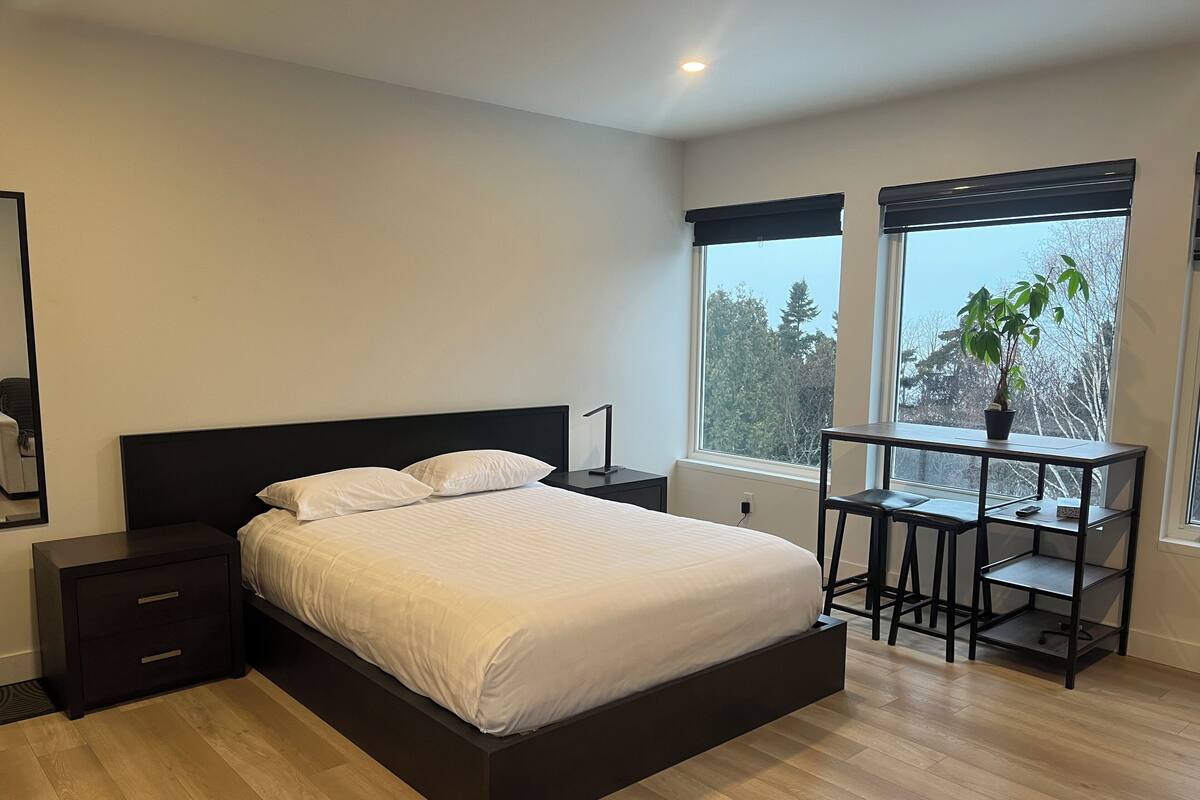 A modern bedroom with a large bed, bedside tables, a desk with a chair, and a window with a view of trees.