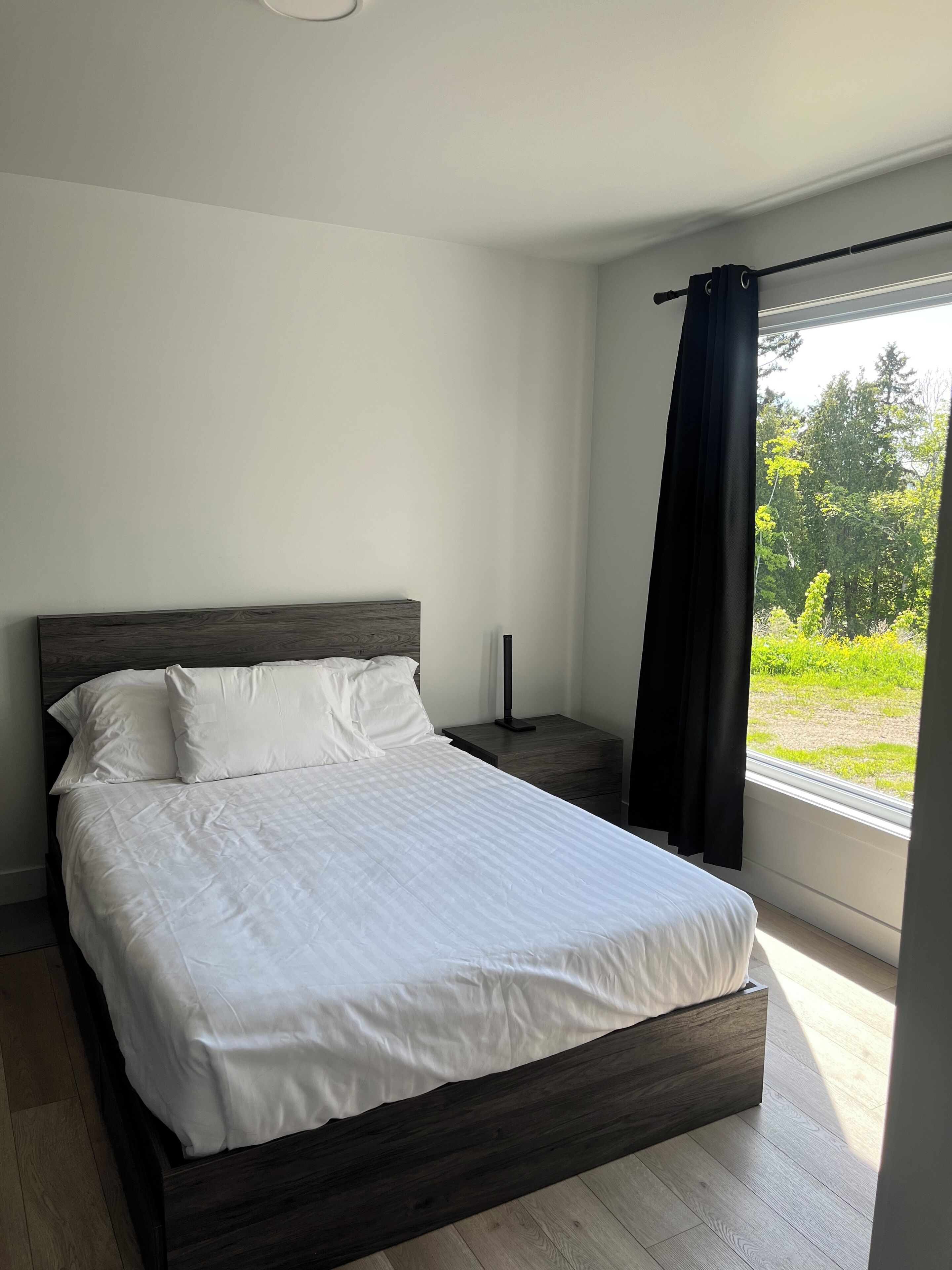Una cama bien hecha con sábanas blancas, un cabecero de madera oscura, una mesita de noche con una lámpara y una ventana con vistas a zonas verdes.