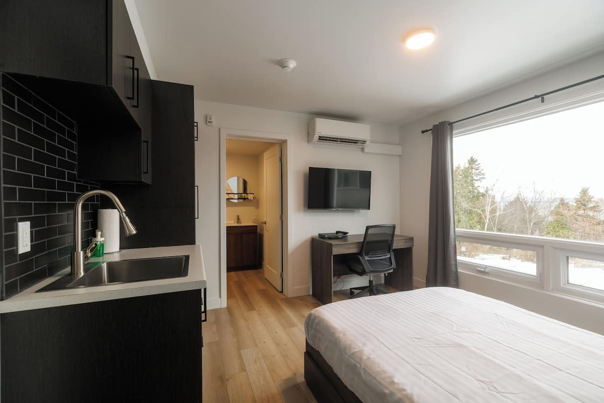 A hotel room with a bed, a desk with a chair, a TV, a kitchen area with a sink, and a window with a view.