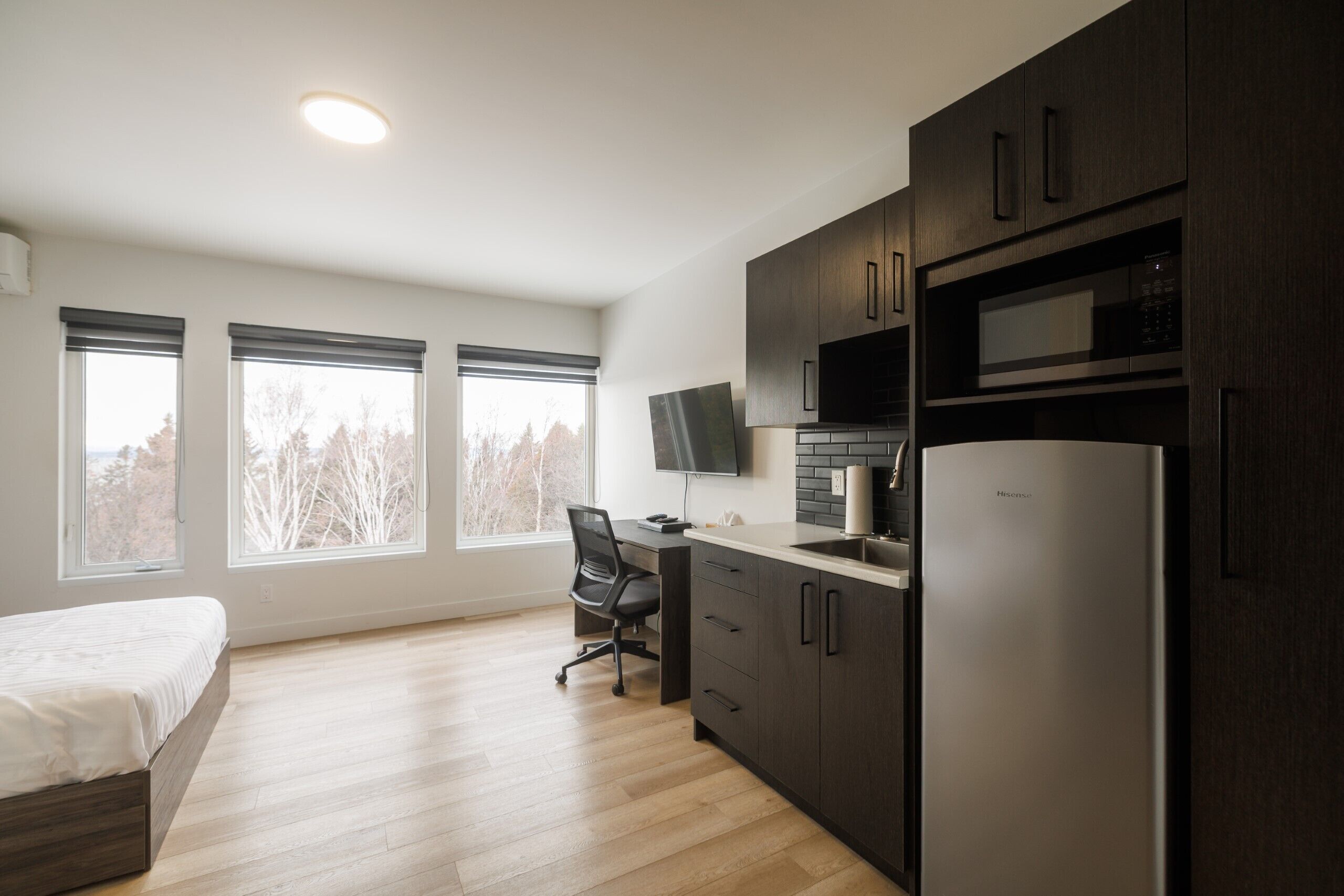 A modern kitchen with dark cabinets, a stainless steel refrigerator, a microwave, and a flat-screen TV. There is a wooden desk with a chair and a bed with a white mattress.
