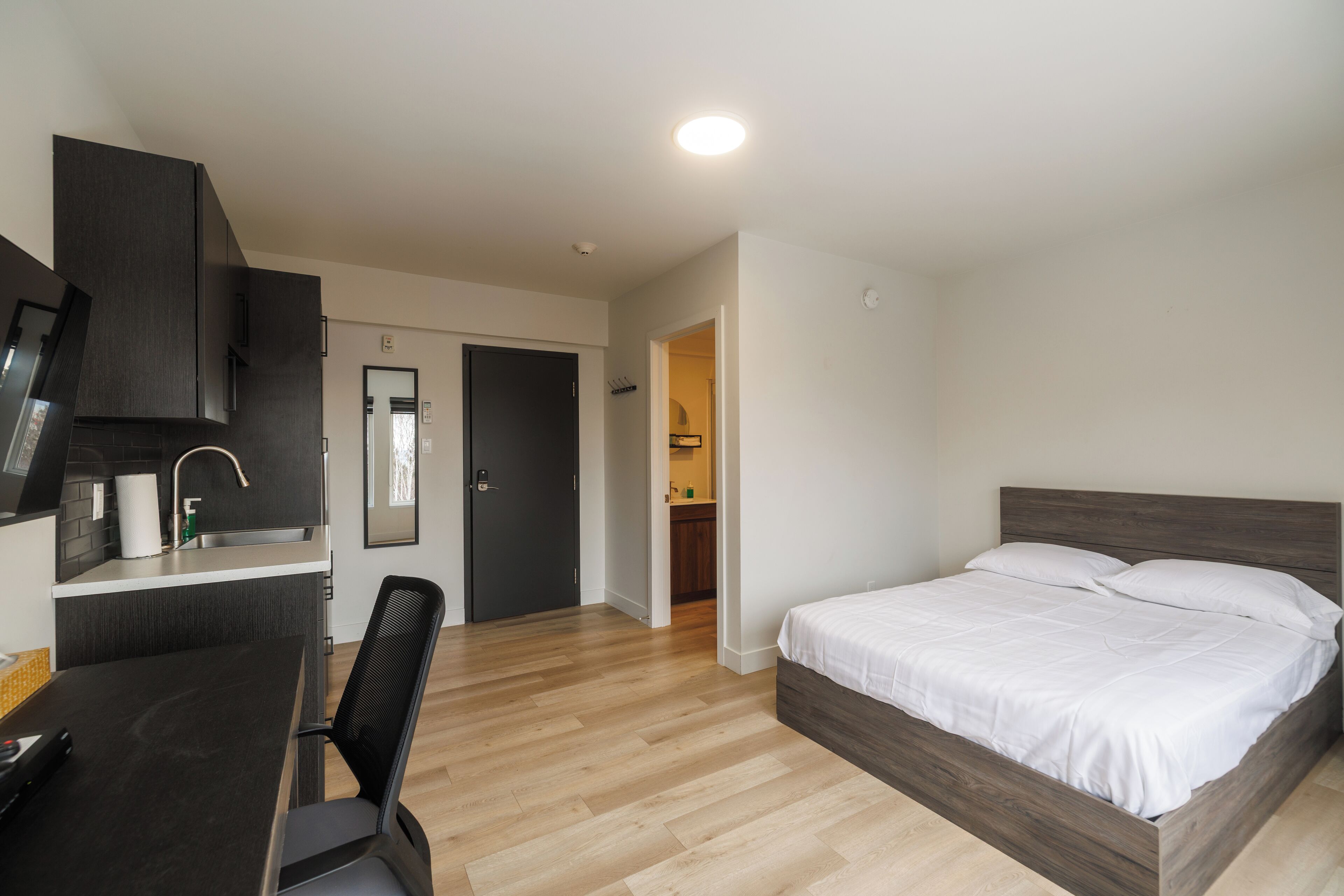 A modern hotel room with a wooden bed, a desk with a chair, a kitchenette, and a bathroom visible through an open door.