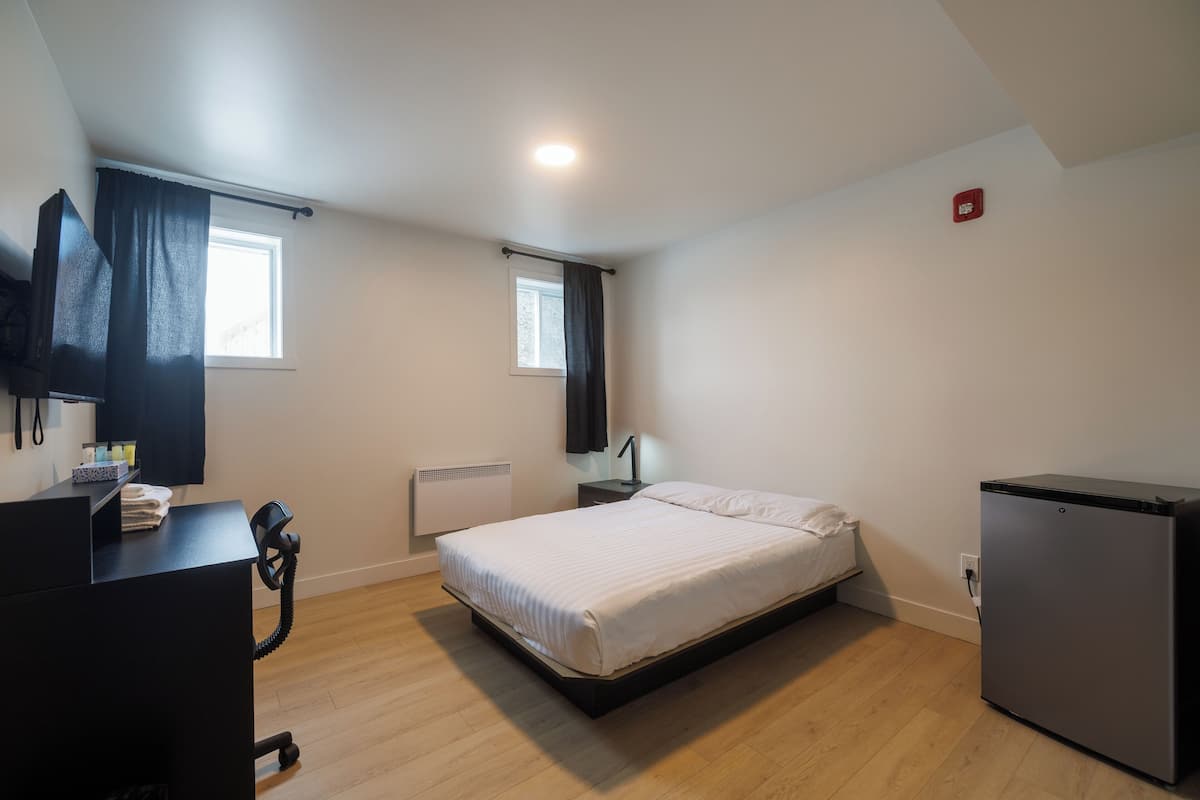 A bedroom with a bed, a desk, a chair, a small refrigerator, and a window.