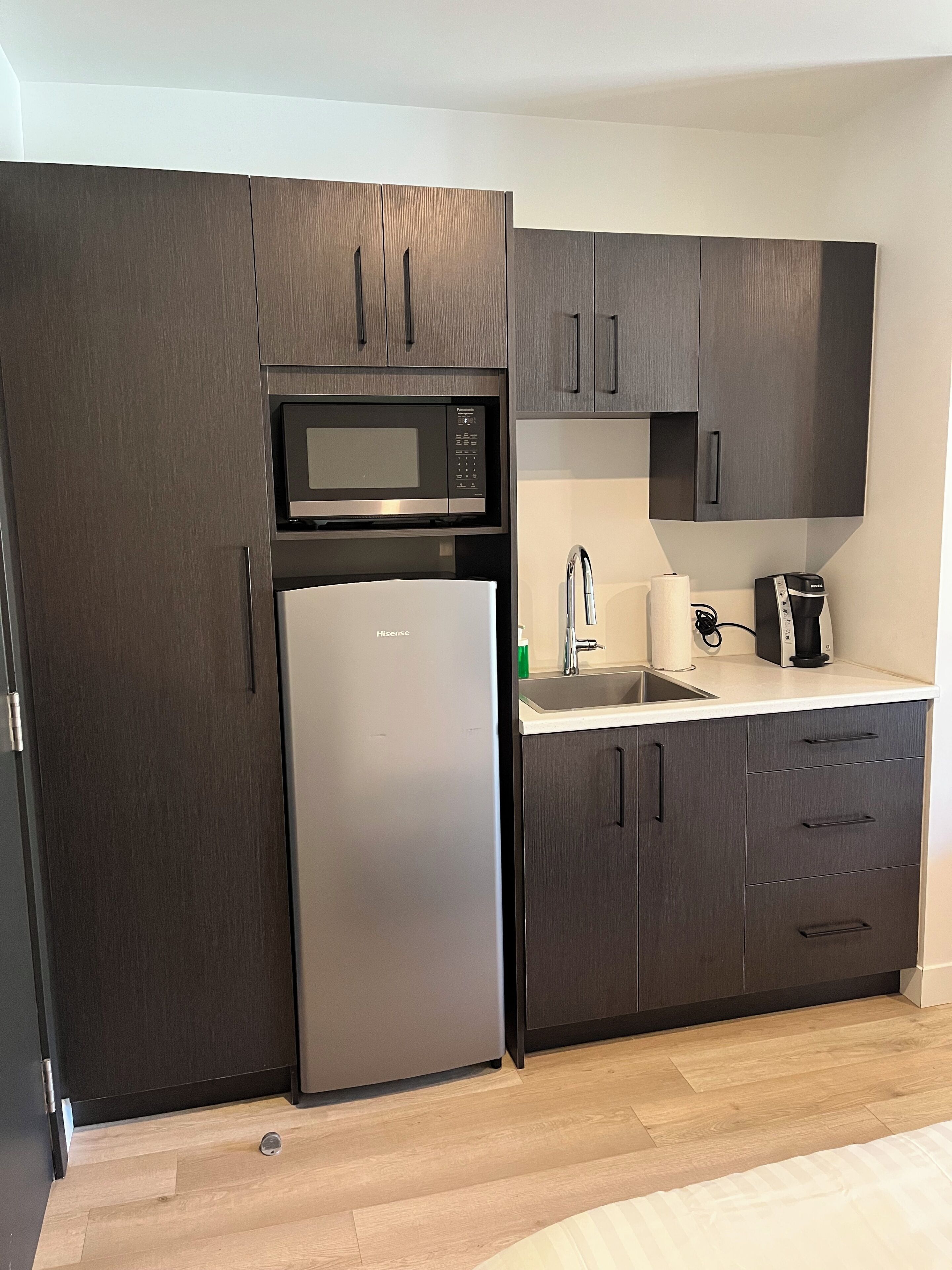 A modern kitchen with dark cabinetry, a stainless steel refrigerator, a microwave, a coffee maker, and a sink.