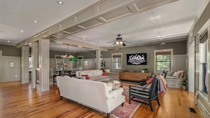 A spacious living room with hardwood floors, a white sofa, a black chair, and a television mounted on the wall. - OBX Rays: Stunning Soundfront Home, Private Heated Pool, Hot Tub, Community Amenities (Corolla)