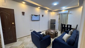 A living room with two sofas, a coffee table, a TV mounted on the wall, a refrigerator, and a door. - Spacious 2-Bedroom Apartment (Hurghada)
