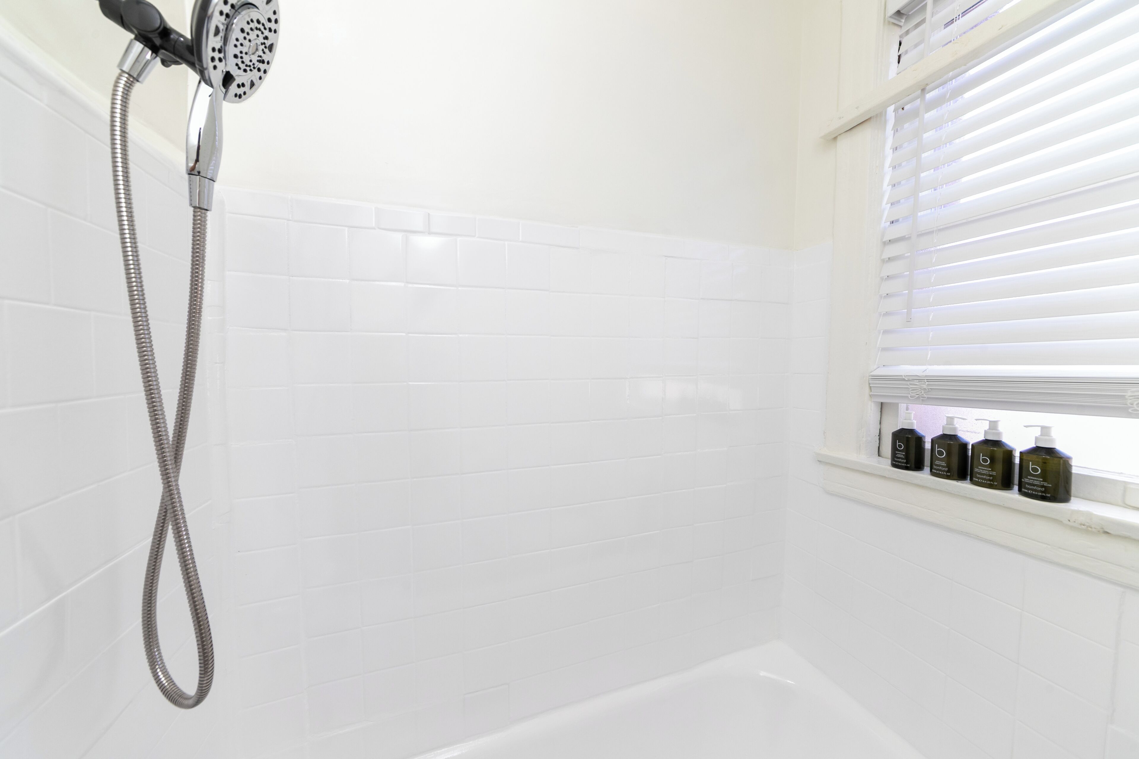 A bathroom with white tiled walls, a shower with a handheld hose, and a window with blinds.