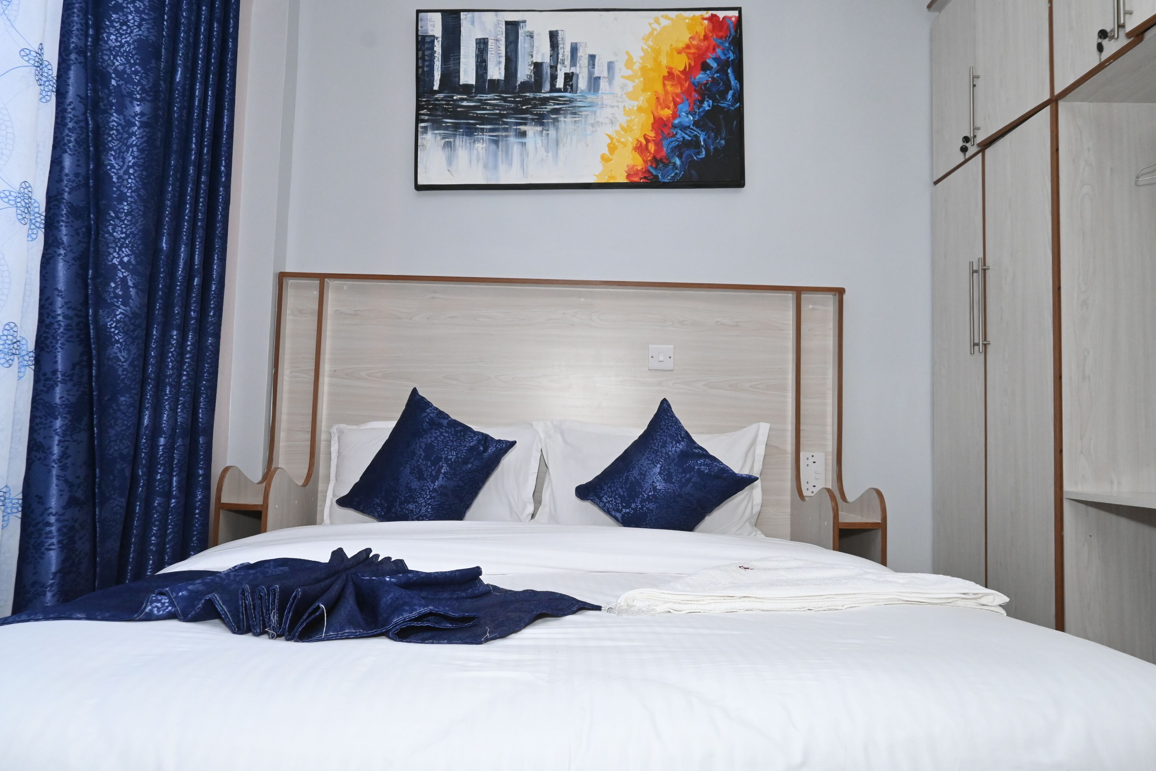 A neatly made bed with white linens and two blue pillows, a cityscape painting above it, and a wardrobe to the right.