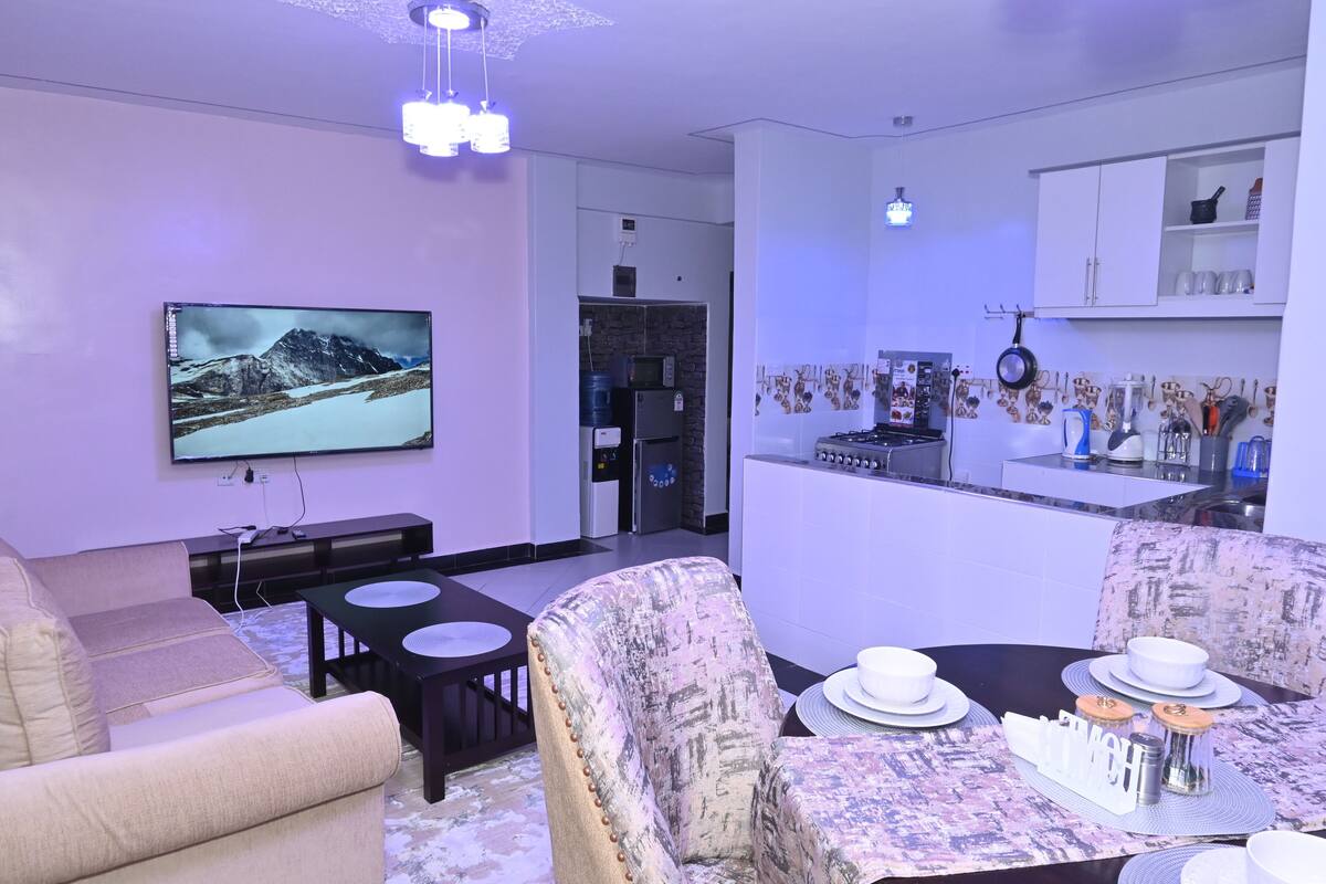 Ein modernes Wohnzimmer mit Flachbildfernseher, einer Couch, einem Esstisch gedeckt mit weißen Tellern und Schüsseln, sowie einer Küchenzeile mit Schränken und einer Mikrowelle.