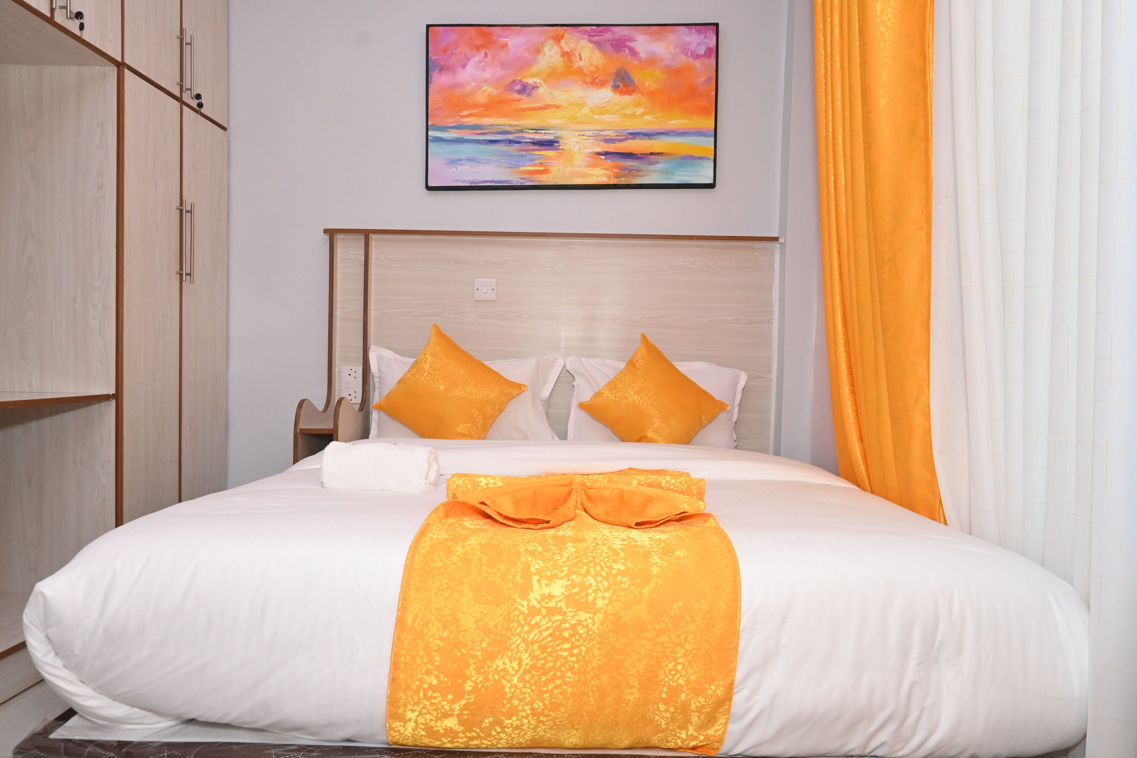A neatly made bed with white and orange bedding, a painting of a sunset on the wall, and a wooden wardrobe to the left.
