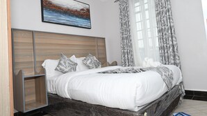 Un dormitorio con cabecera de madera, una cama grande con ropa de cama blanca, un cuadro sobre la cama y una ventana con cortinas estampadas.