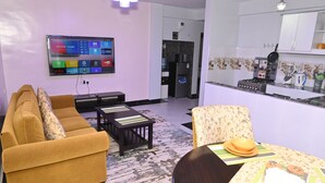 Una sala de estar moderna con un sofá, una mesa de centro y una mesa de comedor preparada para cuatro comensales. Un televisor de pantalla plana está montado en la pared, y se distingue una zona de cocina al fondo.