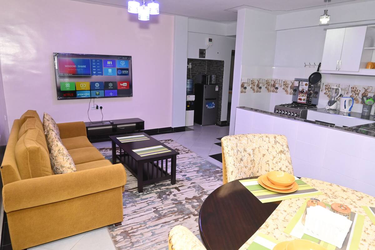 Ein modernes Wohnzimmer mit einer Couch, einem Couchtisch und einem Esstisch für vier Personen. An der Wand ist ein Flachbildfernseher angebracht, und im Hintergrund ist eine Küche erkennbar.