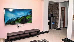 Una sala de estar con un televisor de pantalla plana montado en una pared rosa, un mueble de entretenimiento oscuro y una cocina con protector contra salpicaduras de piedra y electrodomésticos modernos.