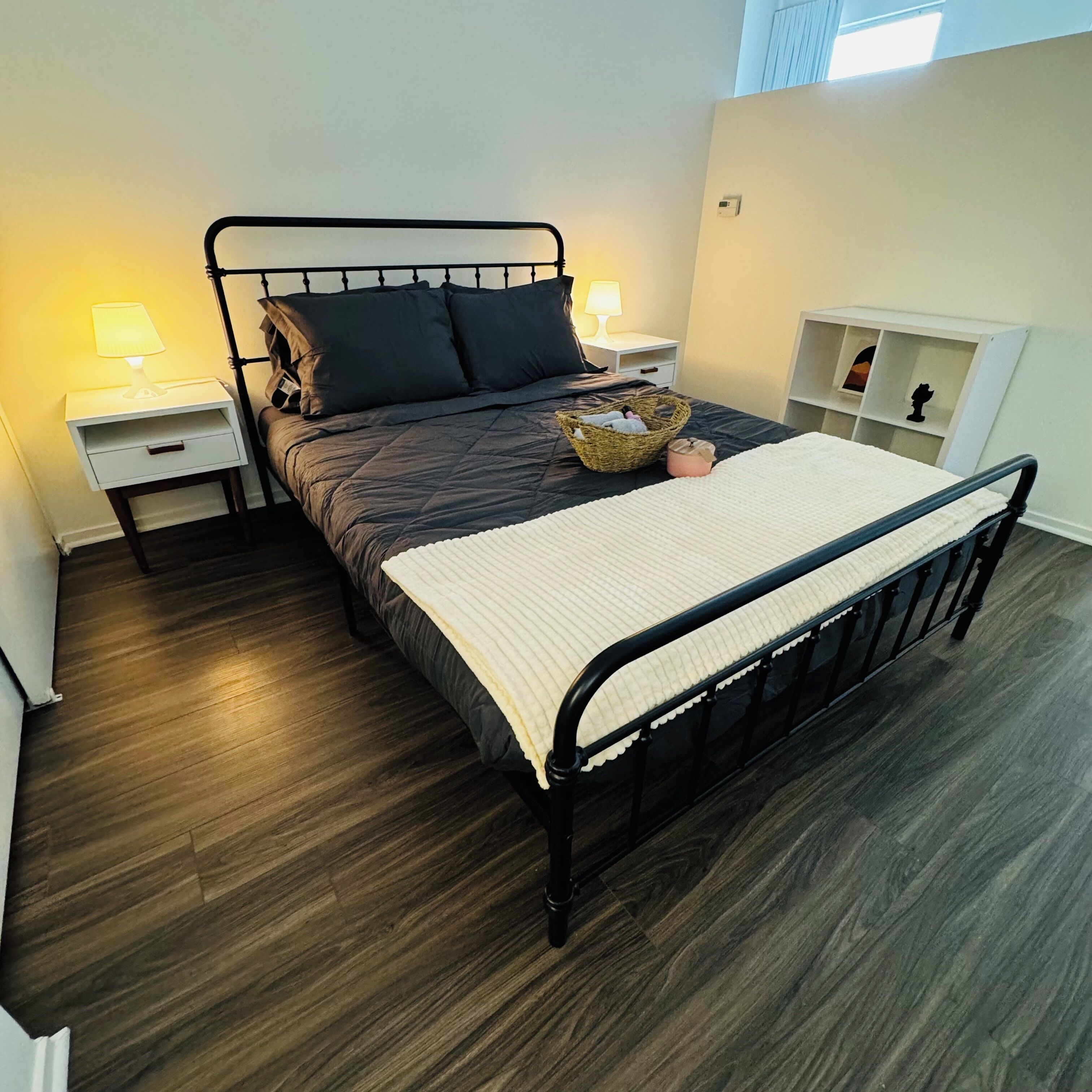 A neatly arranged bedroom with a metal bed frame, dark bedding, a bedside table with a lamp, and a small shelf unit.