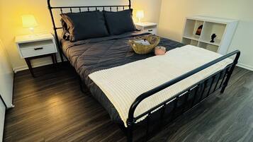 A neatly arranged bedroom with a metal bed frame, dark bedding, a bedside table with a lamp, and a small shelf unit.