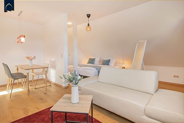 A modern living room with a white sofa, a dining table with chairs, a small coffee table with a vase, and a bed with a nightstand.