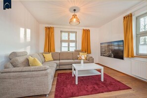 A living room with a sectional sofa, a coffee table, and a red rug. There is a window with yellow curtains and a painting on the wall.