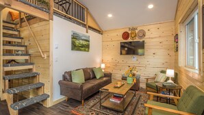 Interior acogedor de una cabaña con escalera de madera, un sofá de cuero marrón, un sillón verde, una mesa de centro de madera y un televisor montado en la pared.