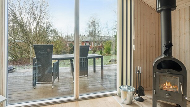 A modern room with a large sliding glass door leading to an outdoor patio, a wood-burning stove, and a dining area with chairs. - Cozy summer house close to the beach (Grenå)
