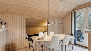 A wooden-paneled room with a round table, white chairs, pendant lights, and a view through a glass door. - Cozy summer house close to the beach (Grenå)
