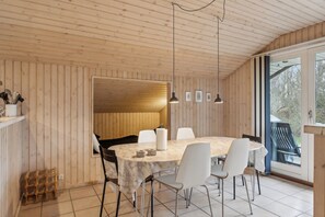 A wooden-paneled room with a round table, white chairs, pendant lights, and a view through a glass door.