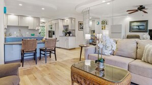 A modern kitchen with white cabinets, a dining area with wicker chairs, a glass coffee table, and a beige sofa.