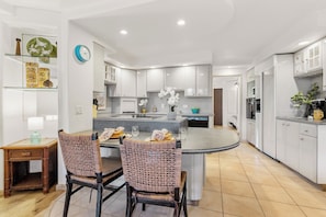 A modern kitchen with white cabinets, a central island, and a dining area with wicker chairs. - Scenic 2BR Retreat Near Maalaea's Pristine Waters (Wailuku)