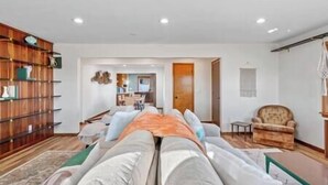 Uma sala de estar com sofá, poltronas, uma estante de madeira e uma área de jantar ao fundo.