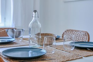 A dining table set with plates, glasses, and a bottle, with a wicker chair and a woven placemat.
