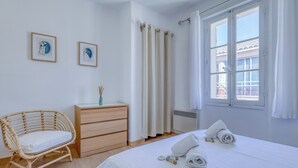 A bedroom with a bed, a wooden dresser, a wicker chair, and two framed artworks on the wall. - Appartement Alegria - Welkeys (Bandol)