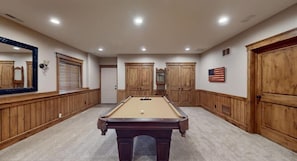 A pool table in a room with wooden paneling and a large mirror on the left.