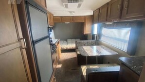 Cocina compacta de autocaravana con armarios de madera, estufa, lavamanos y un pequeño comedor con mesa y banco.