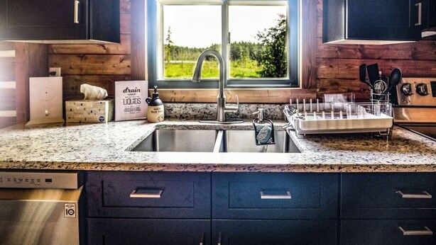 A kitchen with a granite countertop, a double sink, and a window with a view of greenery. - Log Cabin in the Woods (Munising Township)