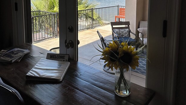 A wooden table with a 'WELCOME' sign, a vase with sunflowers, and a view of a patio with chairs and a palm tree.