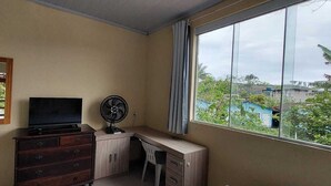 A room with a TV on a wooden dresser, a fan, and a window with a view of greenery and buildings. - Quarto Para Grupo até 4 Pessoas Praia da Maranduba Ubatuba Sp (Sertão da Quina Ubatuba)